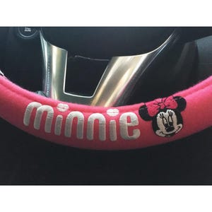 May include: A pink steering wheel cover with the name "Minnie" embroidered in white. The cover features an embroidered image of Minnie Mouse with a pink bow. The steering wheel is silver.