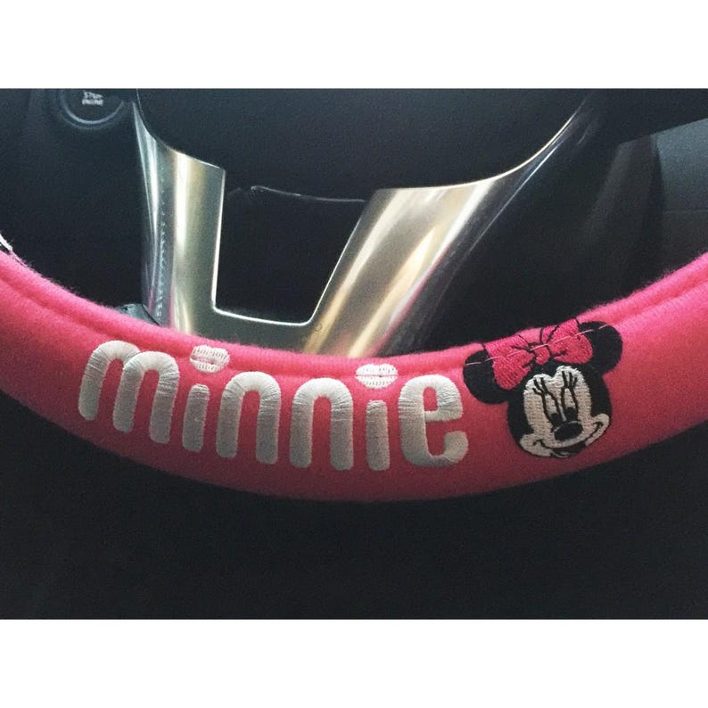 May include: A pink steering wheel cover with the name "Minnie" embroidered in white. The cover features an embroidered image of Minnie Mouse with a pink bow. The steering wheel is silver.