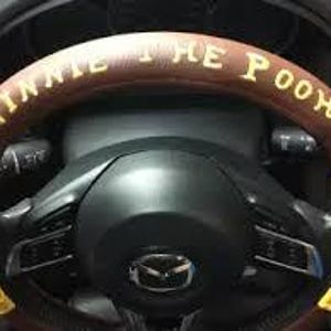 Op de afbeelding: Een bruine stuurwielhoes met de tekst "WINNIE THE POOH" in gele letters. De hoes heeft gele accenten en blauwe patches. Het stuurwiel is zwart met het Mazda-logo.