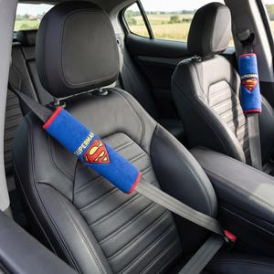 Pode incluir: Duas capas azuis para cinto de segurança com o logótipo vermelho e amarelo do Super-Homem e a palavra SUPERMAN em texto amarelo. As capas têm um rebordo vermelho e estão presas aos cintos cinzentos dos bancos em pele preta.