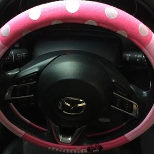 May include: A pink steering wheel cover with white polka dots. The cover is on a black steering wheel with various controls and the Mazda logo. The cover is a car accessory.