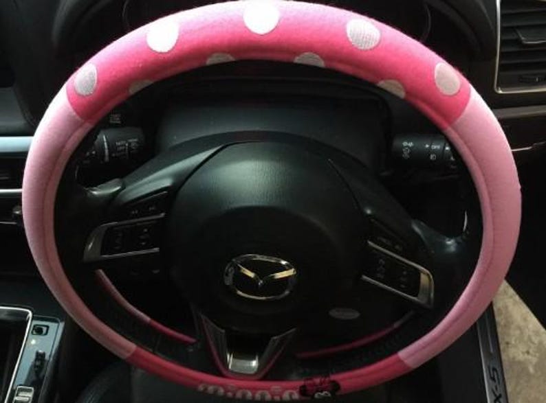 May include: A pink steering wheel cover with white polka dots. The cover is on a black steering wheel with various controls and the Mazda logo. The cover is a car accessory.