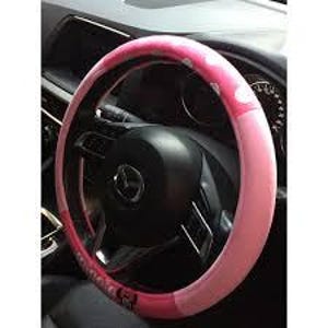 May include: A pink and white polka dot steering wheel cover. The cover is a light pink color with white polka dots and a darker pink band. The cover is on a black steering wheel in a car.