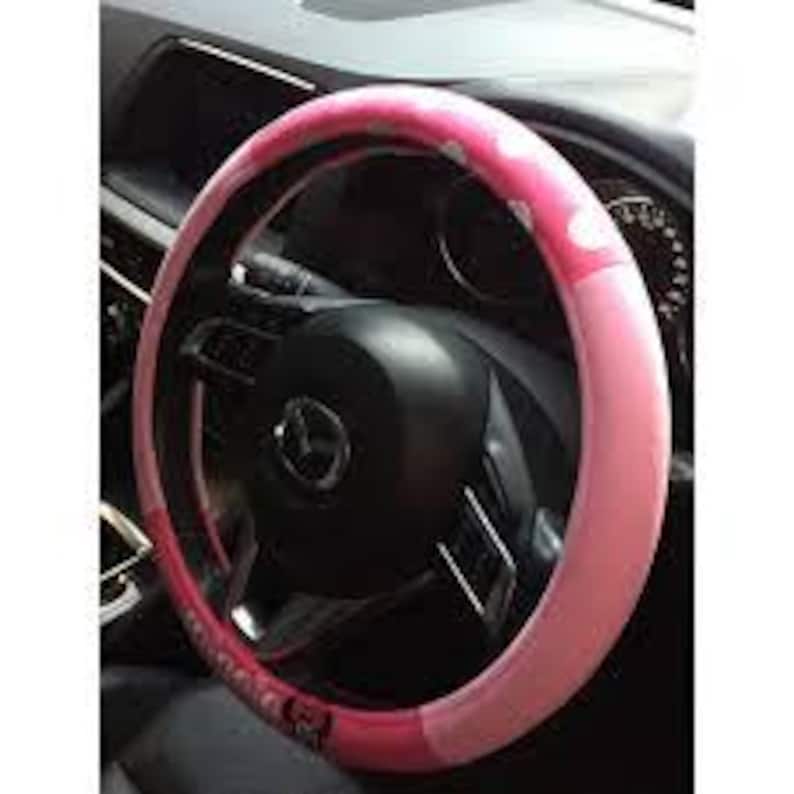 May include: A pink and white polka dot steering wheel cover. The cover is a light pink color with white polka dots and a darker pink band. The cover is on a black steering wheel in a car.