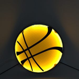 May include: A round, illuminated basketball design with a yellow background and black lines. The basketball is centered and glows against a dark gray background, creating a striking contrast. The design is likely for a sports-themed product.
