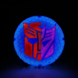 May include: A circular wall sign featuring a split design with a red Autobot logo on the left and a purple Decepticon logo on the right. The background is a blue illuminated honeycomb pattern that glows against the dark, solid black surroundings.