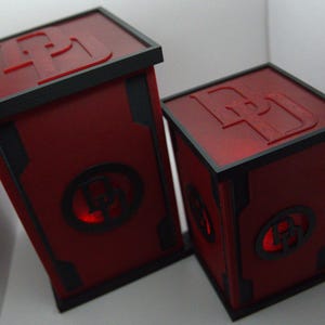 Op de afbeelding: Twee rood-zwarte decoratieve dozen met de letter "D" in reliëf op de boven- en zijkanten. De dozen hebben een vierkante vorm met een zwarte rand en een cirkelvormig ontwerp met de letter "D" op de voorkant. Ze zijn van binnenuit verlicht.