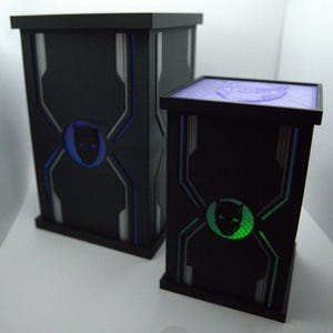 May include: Two black decorative boxes with geometric designs and a panther head emblem. The larger box has a blue emblem, while the smaller box has a green emblem. The tops of the boxes are a light purple colour.