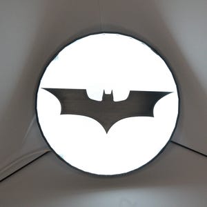 Könnte beinhalten: Ein rundes, beleuchtetes Schild mit dem Batman-Logo. Das Logo ist eine schwarze Silhouette einer Fledermaus vor einem hellweißen Hintergrund. Das Schild ist kreisförmig mit einem schwarzen Rand und an einer grauen Wand befestigt.