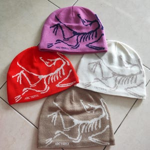 May include: Four beanies in shades of pink, red, cream, and beige. Each beanie displays a white graphic and the brand name "Arc'teryx" in white. The beanies are arranged on a tiled surface.