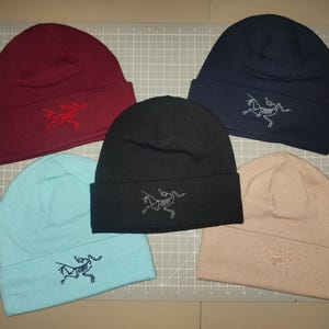 May include: Five beanies in various colors: burgundy, navy blue, black, light blue, and tan. Each beanie has a folded brim and a stitched logo of a skeleton-like creature. The beanies are arranged on a gray cutting mat.
