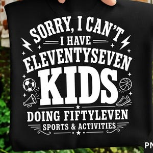 以下が含まれることがあります： 黒いスウェットシャツ。白い文字で「SORRY, I CAN'T - I HAVE ELEVENTYSEVEN KIDS DOING FIFTYLEVEN SPORTS & ACTIVITIES」と書かれています。サッカーボール、バスケットボール、ランニングシューズなどのスポーツ関連のグラフィックが含まれています。