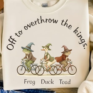 Puede incluir: Sudadera color crema con el texto "Off to overthrow the kings". Presenta una ilustración caprichosa de una rana, un pato y un sapo montando en bicicleta, cada uno con un sombrero de mago y una chaqueta. Las palabras "Frog Duck Toad" están impresas debajo.
