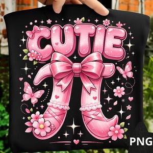 Puede incluir: Camiseta negra con un gráfico rosa. El gráfico presenta la palabra "CUTIE" en letras de burbujas, un gran lazo rosa y piernas rosas con forma de zapatilla de ballet. Mariposas, flores y estrellas rosas completan el diseño.
