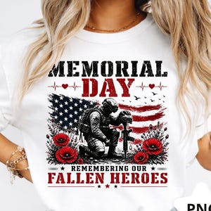 May include: White t-shirt with a Memorial Day graphic. The design includes a kneeling soldier, an American flag, red poppies, and the text "MEMORIAL DAY" and "REMEMBERING OUR FALLEN HEROES".