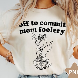 Puede incluir: Camiseta color crema con la frase en negro "off to commit mom foolery". Un gato de dibujos animados con gafas de sol monta en monociclo y sopla burbujas. La imagen tiene una marca de agua PNG en la esquina inferior derecha.