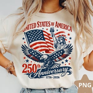 Peut inclure: Sweat-shirt crème avec un motif patriotique. Le graphisme comprend le texte "UNITED STATES OF AMERICA" au-dessus d'un drapeau américain, de la Statue de la Liberté et d'un aigle. Le texte "250th Anniversary 1776-2026" est également affiché.
