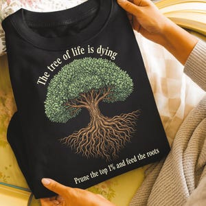 May include: Black sweatshirt featuring a detailed illustration of a tree with a full green canopy and exposed roots. The text "The tree of life is dying" arches above the tree, with "Prune the top 1% and feed the roots" below.