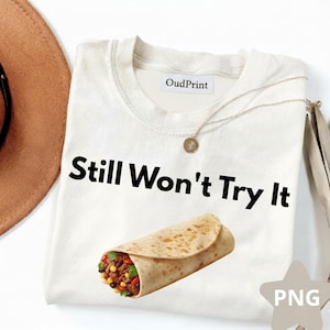 Puede incluir: Camiseta blanca con el texto "Still Won't Try It" en negro. Un gráfico de un burrito está debajo del texto. Un collar dorado y un sombrero marrón son visibles en el fondo.