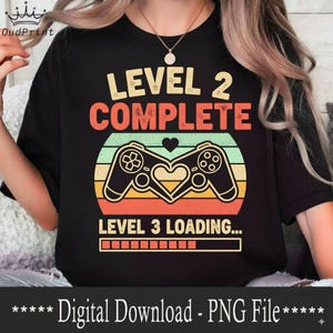 Puede incluir: Camiseta negra con un diseño retro que presenta dos mandos de juego formando un corazón, con el texto "LEVEL 2 COMPLETE" y "LEVEL 3 LOADING...". El diseño tiene un esquema de color vintage.