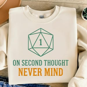 May include: Cream-colored sweatshirt with a green geometric die graphic and the text "ON SECOND THOUGHT NEVER MIND" in green and orange. The sweatshirt is a casual garment.