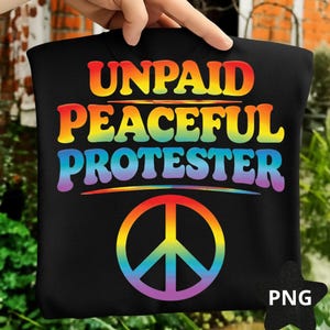 May include: Black t-shirt featuring the text "UNPAID PEACEFUL PROTESTER" in rainbow hues. A rainbow peace symbol is below the text. The shirt is held up by a hand, with a blurred background of greenery and a brick wall.
