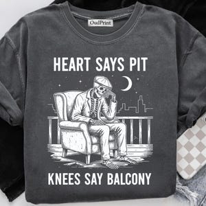 Puede incluir: Sudadera gris oscuro con un gráfico de un esqueleto con traje sentado en un balcón bajo una luna creciente. El texto "HEART SAYS PIT" y "KNEES SAY BALCONY" está impreso encima y debajo del gráfico.