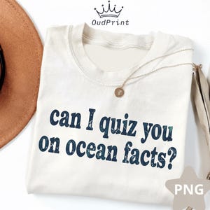 May include: A cream-coloured crew neck t-shirt with the phrase "can I quiz you on ocean facts?" in dark blue text. A gold necklace with a pendant rests on the shirt. A brown hat is visible in the upper left corner.