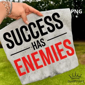 May include: A light gray pillow with the text "SUCCESS HAS ENEMIES" in black and red. The pillow is held by a hand with bracelets. The background is green grass and trees.