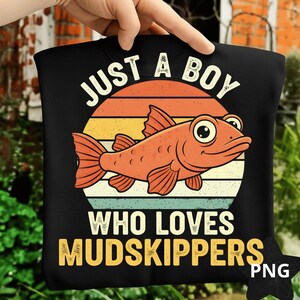 May include: Black t-shirt featuring a cartoon mudskipper fish design. The design includes the text "JUST A BOY WHO LOVES MUDSKIPPERS" in white and gold. The fish is orange and set against a sunset-colored background.