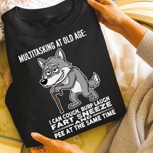 Pu&ograve; includere: Felpa nera con la scritta "MULTITASKING AT OLD AGE: I CAN COUGH, BURP LAUGH FART SNEEZE AND PEE AT THE SAME TIME." Un lupo dei cartoni animati con un bastone &egrave; raffigurato sul davanti.