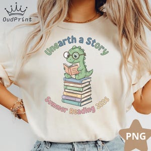 Peut inclure: T-shirt crème avec un dinosaure vert portant des lunettes et lisant un livre sur une pile de livres. Le texte "Unearth a Story" et "Summer Reading 2026" est arqué au-dessus et en dessous de l'illustration du dinosaure.