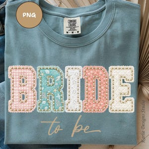 May include: A light blue Comfort Colors t-shirt with the word "BRIDE" in colorful, textured letters. The letters are pink gingham, teal with seashells, white lace, and pink with gold beads. The words "to be" are written below.