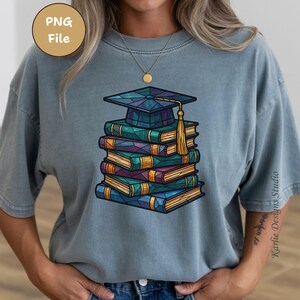 May include: A blue-grey t-shirt featuring a stack of books topped with a graduation cap. The books are in shades of blue, green, and purple, with gold accents. The cap has a gold tassel. The design is a PNG file.