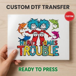 May include: A white DTF transfer featuring the characters Thing 1 and Thing 2 from Dr. Seuss, in red outfits with blue hair. The text "HERE COMES TROUBLE" is in colorful letters, surrounded by cartoon fish. The words "CUSTOM" and "READY TO PRESS" are also visible.