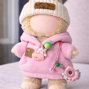 May include: A plush doll wearing a pink hoodie, a white knit hat with bunny ears, and a pacifier necklace. The doll has a small camera charm and a patch that says "Moon Doll".