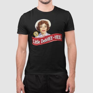 May include: Black t-shirt featuring a graphic of a woman wearing a white hat and a red banner with the text "Little DebHEE-HEE". The graphic includes a gold coin with the text "80s".