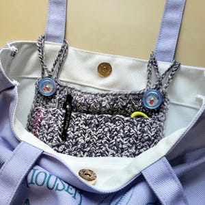 May include: A light purple tote bag with a white interior and a crocheted pocket. The pocket is gray and white with two blue buttons. A black pen and a yellow marker are visible in the pocket. The bag has a magnetic snap closure.