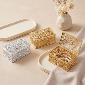 Puede incluir: Tres cajas decorativas de metal en oro y plata. Las cajas tienen intrincados diseños calados. Una caja dorada está abierta, revelando joyas. Un pequeño jarrón blanco con flores está en el fondo.