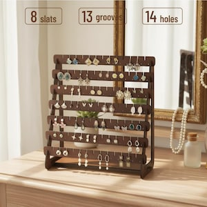 Peut inclure: Un présentoir à boucles d'oreilles en bois marron avec 8 lattes, 13 rainures et 14 trous. Le présentoir est rempli de diverses boucles d'oreilles. Le présentoir est sur une surface en bois.