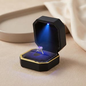 May include: A black octagonal ring box with a gold trim, open to reveal a ring illuminated by a blue light. The ring features a large, round, light-colored stone set in a silver band, resting on a dark blue velvet interior.