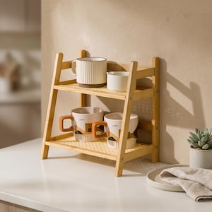 Könnte beinhalten: Ein zweistufiges Bambusregal mit verschiedenen Tassen und einem kleinen weißen Behälter. Die Tassen weisen einzigartige Designs und Farben auf, darunter eine gerippte weiße Tasse mit braunem Boden und Tassen mit braunen Griffen. Das Regal ist aus hellfarbenem Bambus gefertigt.