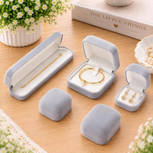 May include: A collection of light gray velvet jewelry boxes, including one long rectangular box containing a gold chain, a square box holding a gold bangle with a pearl, and a small square box displaying a pair of gold flower-shaped earrings.