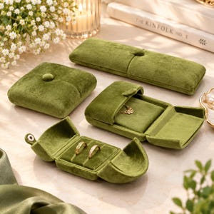 May include: A collection of olive green velvet jewelry boxes in various shapes and sizes. One rectangular box is closed, one square box is closed, one rectangular box is open holding a gold necklace, and one small oval box is open holding two rings.