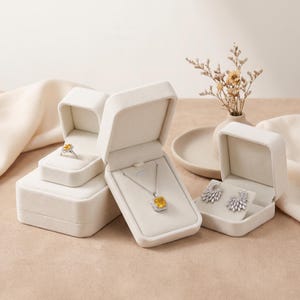 May include: A set of three cream-colored velvet jewelry boxes containing a silver ring with a yellow gemstone, a silver necklace with a rectangular yellow pendant, and a pair of silver earrings featuring marquise-cut clear stones.