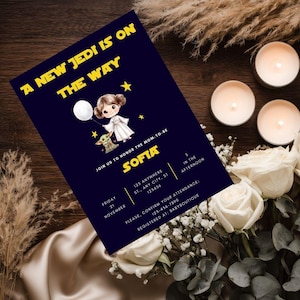 Star Wars baby shower invitation displayed in a white frame on a wooden table, featuring chibi Princess Leia holding a balloon and Baby Yoda on a navy galaxy background with gold text 'A New Jedi is on the Way'.
