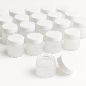 Puede incluir: Un conjunto de pequeños frascos de vidrio esmerilado con tapas blancas. Un frasco está abierto, con su tapa al lado. Los frascos están dispuestos sobre una superficie blanca, lo que sugiere un producto para almacenar cremas, bálsamos u otros artículos pequeños. La estética general es limpia y minimalista.