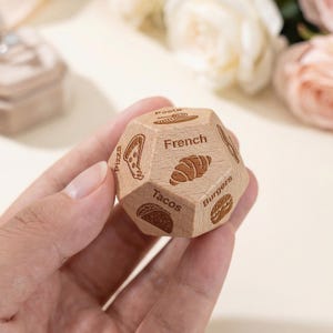 May include: A wooden die with multiple sides, each engraved with a food illustration and label. The die shows pizza, tacos, burgers, pasta, and a croissant, with the word "French" on one side. The die is held in a hand.