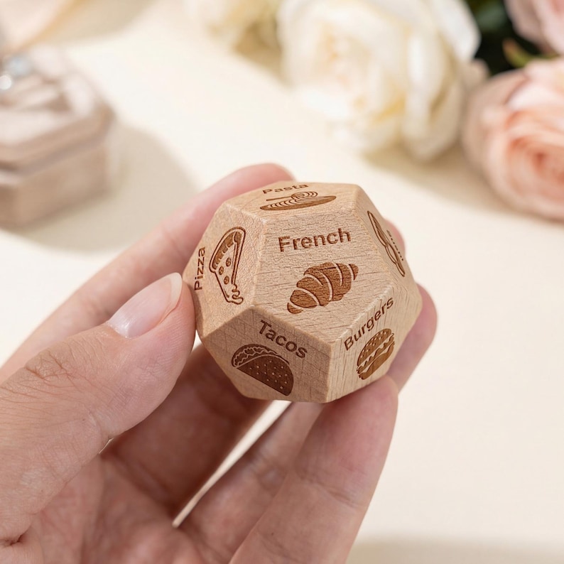 May include: A wooden die with multiple sides, each engraved with a food illustration and label. The die shows pizza, tacos, burgers, pasta, and a croissant, with the word "French" on one side. The die is held in a hand.