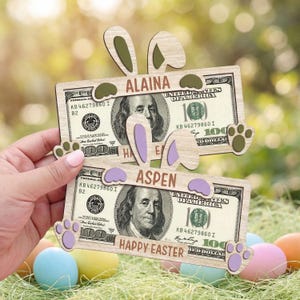 May include: Wooden Easter bunny ear and paw decorations with the names "ALAINA" and "ASPEN" on them, placed over two one-hundred-dollar bills. The words "HAPPY EASTER" are also visible. The image is set against a blurred background with Easter eggs.